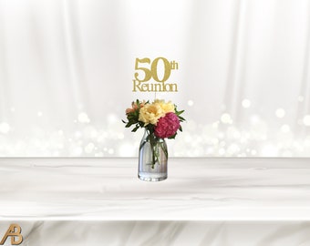 May include: Gold glitter 50th reunion centerpiece with the words "50th Reunion" on a stick. The stick is inserted into a clear glass vase with pink, yellow, and white flowers.