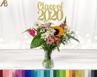 May include: A clear glass vase holds a vibrant bouquet of flowers, including a sunflower, daisies, and pink blossoms. A gold glitter "Class of 2020" topper sits above the flowers. The background is a soft white with bokeh.