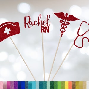 May include: Four red glitter cake toppers with a nurse's hat, the name Rachel RN, a caduceus, and a stethoscope. The toppers are on wooden sticks.