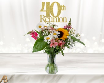 May include: Gold glitter 40th Reunion cake topper with the word "Reunion" below the number 40. The topper is placed on a bouquet of flowers in a clear glass vase.