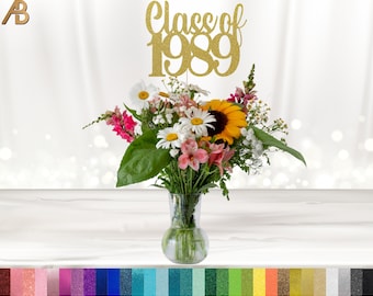May include: Gold glitter Class of 1989 topper for a graduation centerpiece. The topper is placed on a bouquet of flowers in a clear glass vase.