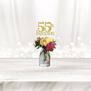 May include: Gold glitter 55th Reunion centerpiece with the text "55th Reunion" on a white background. A clear glass vase with pink and yellow flowers sits on a white table.