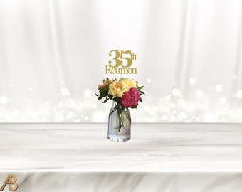 May include: Gold glitter 35th reunion centerpiece with the text "35th Reunion" on top of a clear glass vase with pink, white, and yellow flowers.