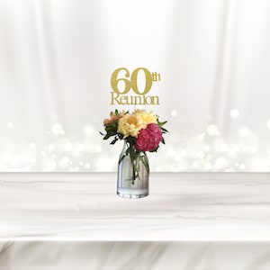 May include: Gold glitter 60th reunion centerpiece with the text "60th Reunion" and a vase of pink, white, and peach flowers.