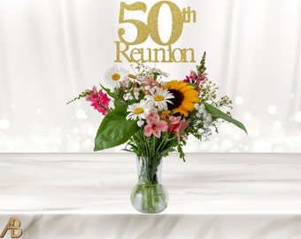 May include: A bouquet of flowers in a clear glass vase with a gold glitter 50th Reunion topper. The flowers include white daisies, pink alstroemeria, yellow sunflowers, and green foliage.