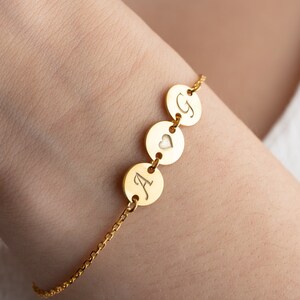 May include: A gold chain bracelet with three round charms. The charms are engraved with the letters "A", a heart, and "G".