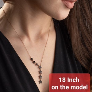 May include: A necklace featuring a series of star pendants. The necklace is 45.7 cm long and has a cascade of star-shaped charms. The stars are adorned with dark accents, creating a striking contrast against the chain. The necklace is worn against a black top.