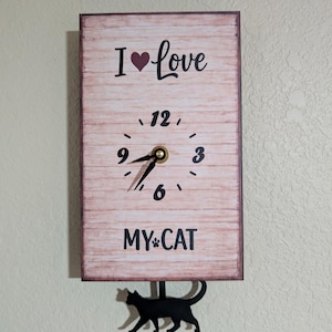 May include: A rectangular wall clock with a light brown wood grain background. The clock face has black numbers and hands, with the text "I love MY CAT". A black cat silhouette hangs below the clock.