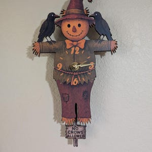 May include: A wooden scarecrow clock with a smiling face, wearing a hat and bow tie. Two black crows perch on its shoulders. The clock hands point to 12 and 16. A sign below reads "NO CROWS ALLOWED."