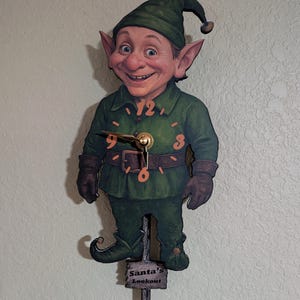 May include: A whimsical wall clock shaped like a smiling elf, dressed in a green outfit with a matching hat. The clock face features large orange numbers and gold hands. The words "Santa's Lookout" are printed on a small sign at the bottom.