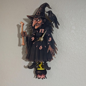 May include: A whimsical Halloween-themed clock featuring a cartoon witch with a long nose, holding a bone, and wearing a black hat and dress. A black crow sits on her shoulder. The clock hands are gold, and the numbers are pink. The witch stands above a cauldron.