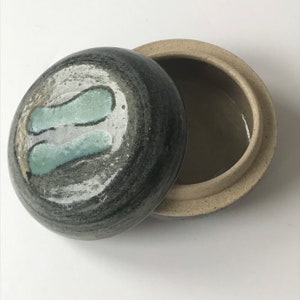 Pode incluir: Uma caixa redonda de cerâmica para miudezas com tampa. A tampa é decorada com um padrão de espiral preto e branco e um design azul claro. A caixa é feita de cerâmica marrom claro.