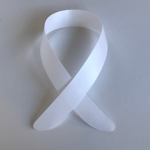Puede incluir: Una cinta de raso blanca, doblada en un lazo, formando una cinta de concienciación simbólica. La cinta es lisa y tiene un ligero brillo, con los extremos cuidadosamente metidos. La cinta se muestra sobre un fondo blanco liso.