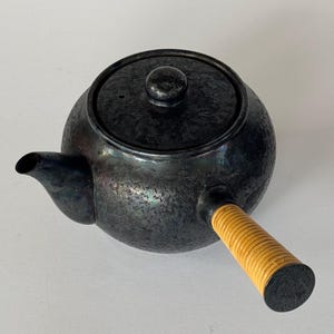 May include: Black teapot with a textured surface and a wooden handle. The teapot has a lid with a small knob on top.
