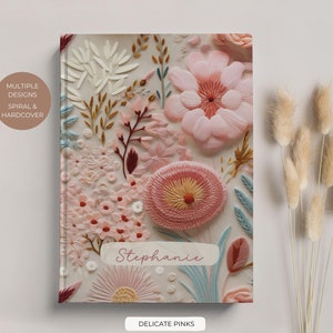 May include: A hardcover journal with a floral design featuring delicate pink and white flowers. The journal has the name "Stephanie" written on the front and the text "Delicate Pinks" at the bottom.