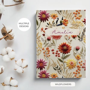 K&ouml;nnte beinhalten: Ein personalisiertes Notizbuch mit einem floralen Design mit gestickten Wildblumen in Rot-, Orange-, Gelb- und Rosat&ouml;nen. Der Notizbuchumschlag tr&auml;gt den Namen "Amelia" und die Worte "Wildflowers" unten.