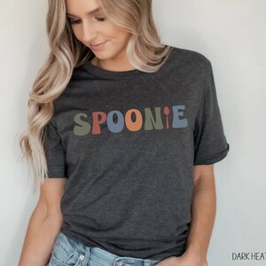 K&ouml;nnte beinhalten: Dunkelgraues T-Shirt mit dem Wort "SPOONIE" in bunten, Retro-Lettern. Das Wort "SPOONIE" ist in einer verspielten Schriftart mit einer L&ouml;ffelgrafik am Ende.