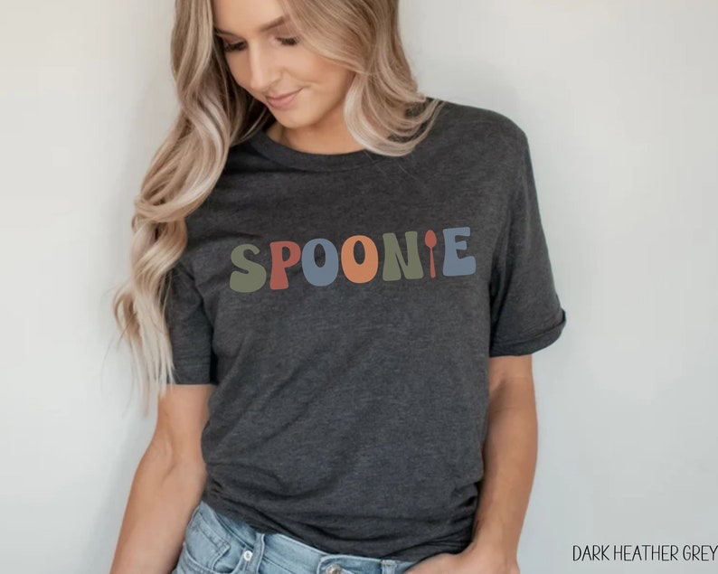 K&ouml;nnte beinhalten: Dunkelgraues T-Shirt mit dem Wort "SPOONIE" in bunten, Retro-Lettern. Das Wort "SPOONIE" ist in einer verspielten Schriftart mit einer L&ouml;ffelgrafik am Ende.