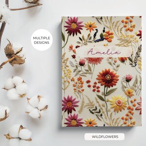 May include: A cream-coloured notebook cover with a floral design featuring embroidered wildflowers in shades of red, orange, yellow, and pink. The name "Amelia" is written in cursive on a white banner.