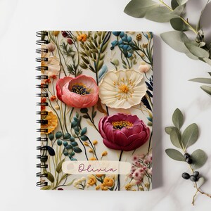 K&ouml;nnte beinhalten: Ein spiralgebundenes Notizbuch mit einem Blumendesign mit rosa, roten und wei&szlig;en Blumen. Der Einband tr&auml;gt den Namen "Olivia" aufgedruckt.