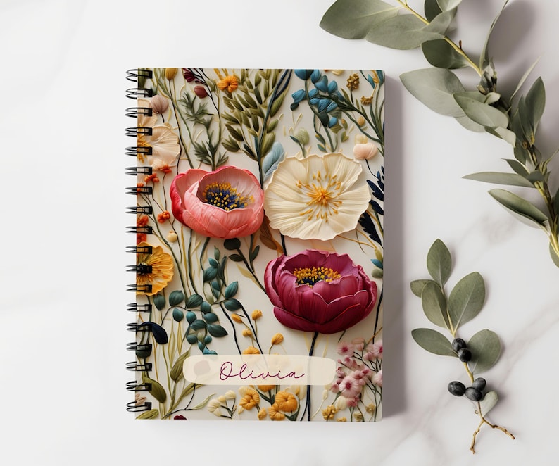 K&ouml;nnte beinhalten: Ein spiralgebundenes Notizbuch mit einem Blumendesign mit rosa, roten und wei&szlig;en Blumen. Der Einband tr&auml;gt den Namen "Olivia" aufgedruckt.