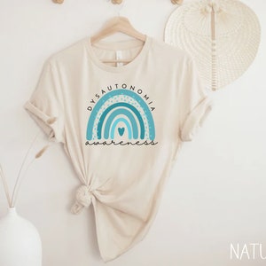 Puede incluir: Una camiseta beige con un arco&iacute;ris azul y turquesa y el texto "Dysautonomia Awareness".