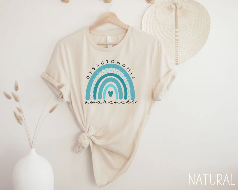 Puede incluir: Una camiseta beige con un arco&iacute;ris azul y turquesa y el texto "Dysautonomia Awareness".