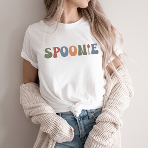 K&ouml;nnte beinhalten: Wei&szlig;es T-Shirt mit dem Wort "SPOONIE" in bunten, abgerundeten Buchstaben. Das "I" wird durch einen L&ouml;ffel ersetzt.