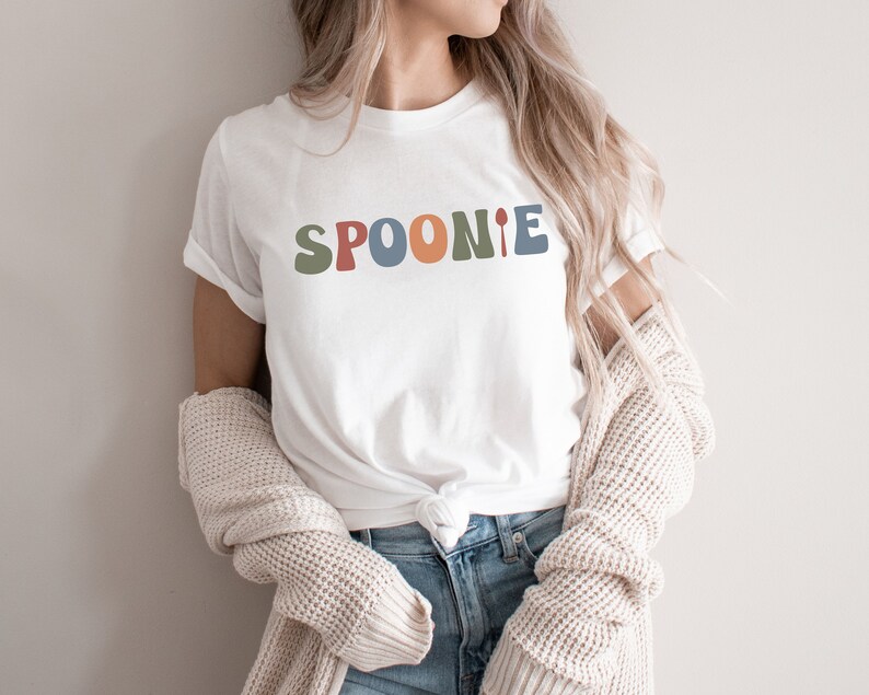 K&ouml;nnte beinhalten: Wei&szlig;es T-Shirt mit dem Wort "SPOONIE" in bunten, abgerundeten Buchstaben. Das "I" wird durch einen L&ouml;ffel ersetzt.