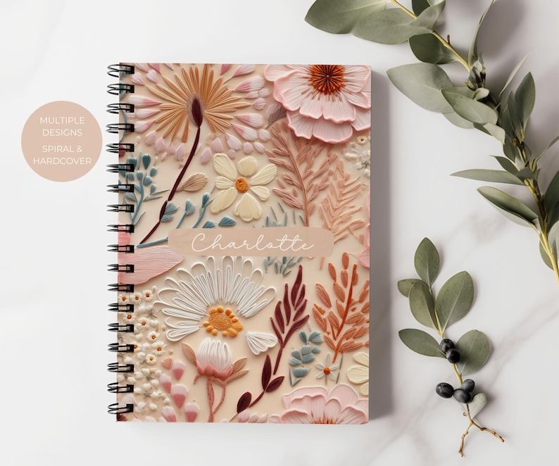 Puede incluir: Un cuaderno de tapa dura con espiral y un dise&ntilde;o floral. La portada presenta una variedad de flores en tonos rosa, blanco y marr&oacute;n. El nombre "Charlotte" est&aacute; impreso en la portada.