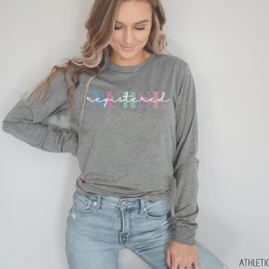 May include: A gray long-sleeve shirt with the text "registered nurse" in colorful letters. The shirt is being worn by a person with long brown hair.