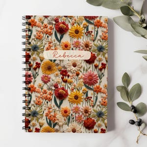 K&ouml;nnte beinhalten: Ein spiralgebundenes Notizbuch mit einem Blumenmuster. Der Einband zeigt eine Vielzahl von bunten Blumen, darunter rote, gelbe, rosa und orangefarbene. Der Name "Rebecca" ist auf dem Einband aufgedruckt.