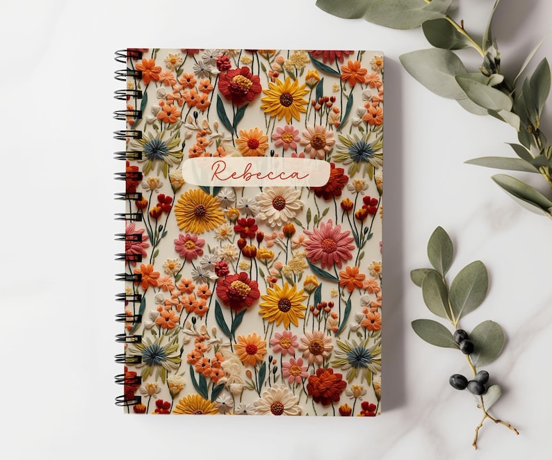 K&ouml;nnte beinhalten: Ein spiralgebundenes Notizbuch mit einem Blumenmuster. Der Einband zeigt eine Vielzahl von bunten Blumen, darunter rote, gelbe, rosa und orangefarbene. Der Name "Rebecca" ist auf dem Einband aufgedruckt.