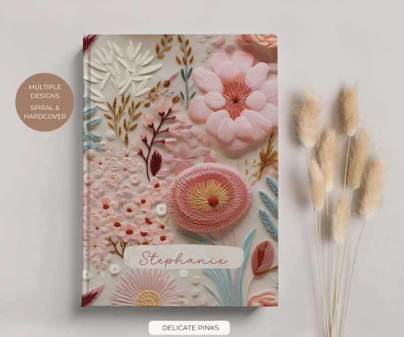 May include: A hardcover journal with a floral design featuring delicate pink and white flowers. The journal has the name "Stephanie" written on the front and the text "Delicate Pinks" at the bottom.