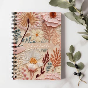 May include: A spiral bound notebook with a floral design. The cover features a pink, white and brown floral pattern with the name "Charlotte" printed on the front.