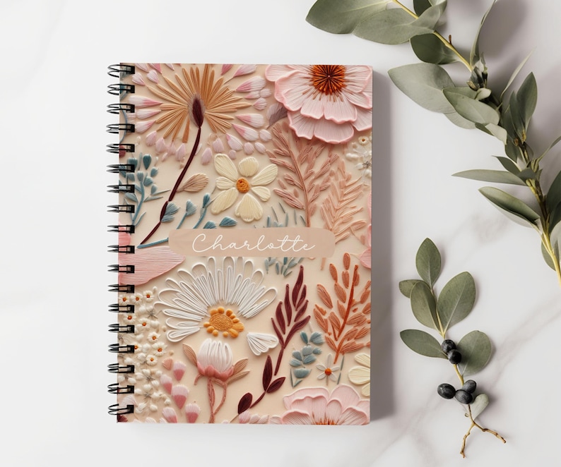 K&ouml;nnte beinhalten: Ein spiralgebundenes Notizbuch mit einem floralen Design. Der Einband zeigt ein pinkfarbenes, wei&szlig;es und braunes Blumenmuster mit dem Namen "Charlotte" auf der Vorderseite.