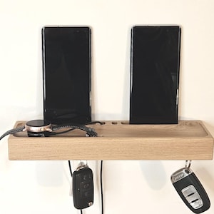 Può includere: Una stazione di ricarica in legno di rovere chiaro con due smartphone neri, uno smartwatch e due mazzi di chiavi. La stazione ha un ripiano con fessure per dispositivi e ganci per chiavi sottostanti. L'orologio ha un cinturino in pelle nera.