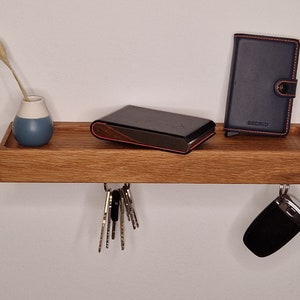 Può includere: Un ripiano in legno a parete con ganci per le chiavi. Il ripiano ha un piccolo vaso blu con un fiore secco, un portafoglio nero con la scritta "Secrid" e un oggetto rettangolare nero in cima.