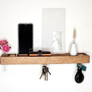 K&ouml;nnte beinhalten: Ein h&ouml;lzernes Wandregal mit verschiedenen Gegenst&auml;nden. Ein Smartphone, eine Geldb&ouml;rse und Schl&uuml;ssel befinden sich auf dem Regal. Eine kleine Vase mit einer getrockneten Pflanze und eine mit rosa Blumen sind ebenfalls vorhanden. Das Regal ist an einer wei&szlig;en Wand befestigt.