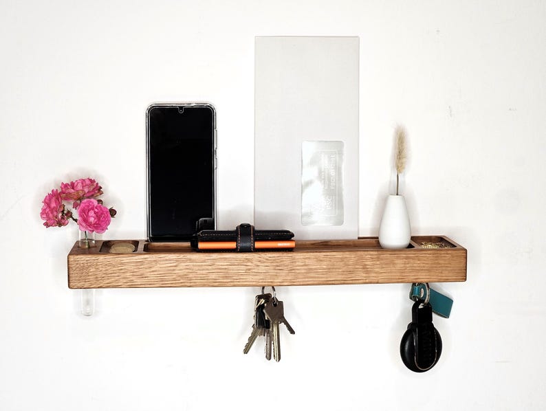 K&ouml;nnte beinhalten: Ein h&ouml;lzernes Wandregal mit verschiedenen Gegenst&auml;nden. Ein Smartphone, eine Geldb&ouml;rse und Schl&uuml;ssel befinden sich auf dem Regal. Eine kleine Vase mit einer getrockneten Pflanze und eine mit rosa Blumen sind ebenfalls vorhanden. Das Regal ist an einer wei&szlig;en Wand befestigt.