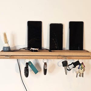 Può includere: Una stazione di ricarica in legno con tre smartphone neri, un piccolo vaso con erba secca e un orologio. Sotto, ci sono ganci con chiavi e portachiavi. La stazione è montata su un muro bianco.