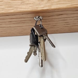 K&ouml;nnte beinhalten: Ein h&ouml;lzerner Schl&uuml;sselhalter mit einem Satz Schl&uuml;ssel, die an einem Metallhaken h&auml;ngen. Der Schl&uuml;sselhalter ist aus hellfarbenem Holz gefertigt und hat zwei wei&szlig;e kreisf&ouml;rmige Magnete. Die Schl&uuml;ssel sind in verschiedenen Gr&ouml;&szlig;en und Formen.
