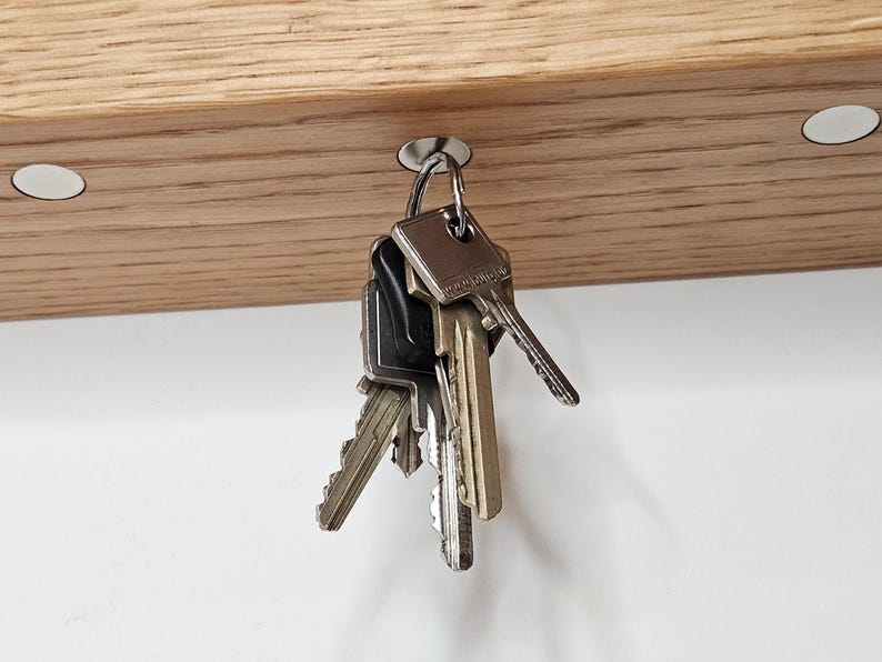K&ouml;nnte beinhalten: Ein h&ouml;lzerner Schl&uuml;sselhalter mit einem Satz Schl&uuml;ssel, die an einem Metallhaken h&auml;ngen. Der Schl&uuml;sselhalter ist aus hellfarbenem Holz gefertigt und hat zwei wei&szlig;e kreisf&ouml;rmige Magnete. Die Schl&uuml;ssel sind in verschiedenen Gr&ouml;&szlig;en und Formen.