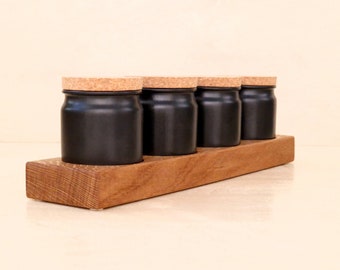 Op de afbeelding: Vier zwarte specerijpotten met kurken deksels op een houten rek. De potten zijn in een rij op het rek geplaatst, dat van lichtbruin hout is gemaakt.
