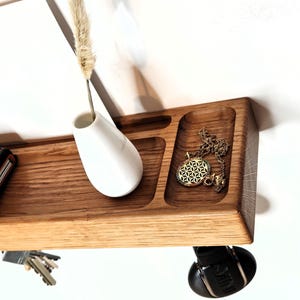 K&ouml;nnte beinhalten: Ein schwebendes Holzregal mit integrierter Ablage, das eine wei&szlig;e Vase mit einer getrockneten Pflanze, eine goldene Halskette, eine Brieftasche und Schl&uuml;ssel enth&auml;lt. Das Regal ist an einer wei&szlig;en Wand befestigt und schafft eine minimalistische &Auml;sthetik.