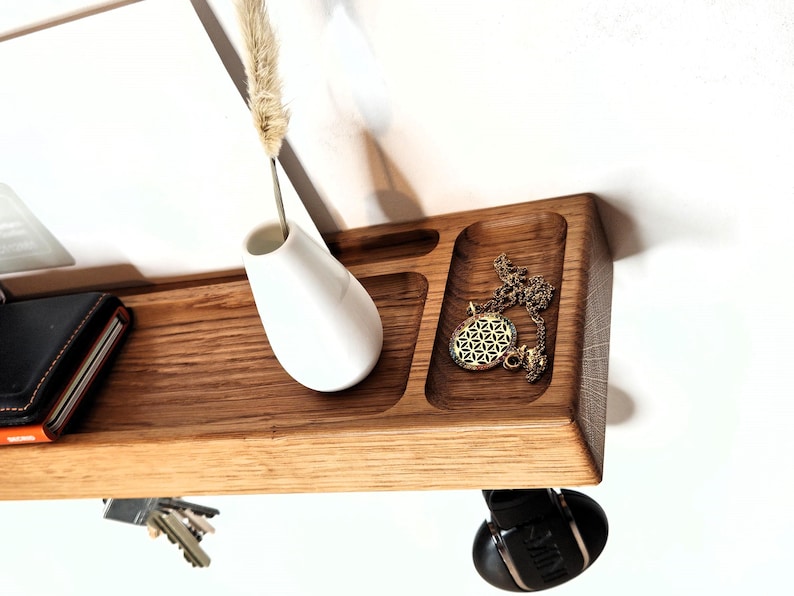 K&ouml;nnte beinhalten: Ein schwebendes Holzregal mit integrierter Ablage, das eine wei&szlig;e Vase mit einer getrockneten Pflanze, eine goldene Halskette, eine Brieftasche und Schl&uuml;ssel enth&auml;lt. Das Regal ist an einer wei&szlig;en Wand befestigt und schafft eine minimalistische &Auml;sthetik.