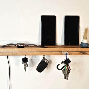 Könnte beinhalten: Ein Wandorganizer aus Holz mit einem Regal für Telefone, eine Uhr und eine kleine Vase. Darunter befinden sich Haken für Schlüssel und einen Autoschlüsselanhänger. Ein schwarzes Ladekabel hängt herunter. Der Organizer ist aus hellem Holz.