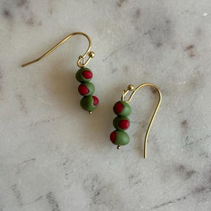 May include: A pair of gold-colored dangle earrings. Each earring features three olive-shaped beads in green with a red center, strung on a gold-colored wire. The earrings are displayed on a white and gray marble surface.