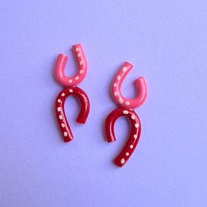 May include: A pair of handmade, abstract earrings. Each earring features a curved, horseshoe-like shape in two stacked sections. The top section is pink with white dots, and the bottom is red with white dots. The earrings are set against a lavender background.