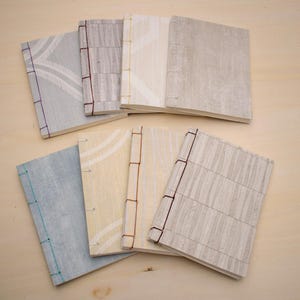 May include: A collection of handmade journals with textured covers in various colors, including light blue, beige, and gray. Each journal is bound with visible stitching in coordinating colors like purple, brown, and green. The journals are arranged on a light wooden surface.